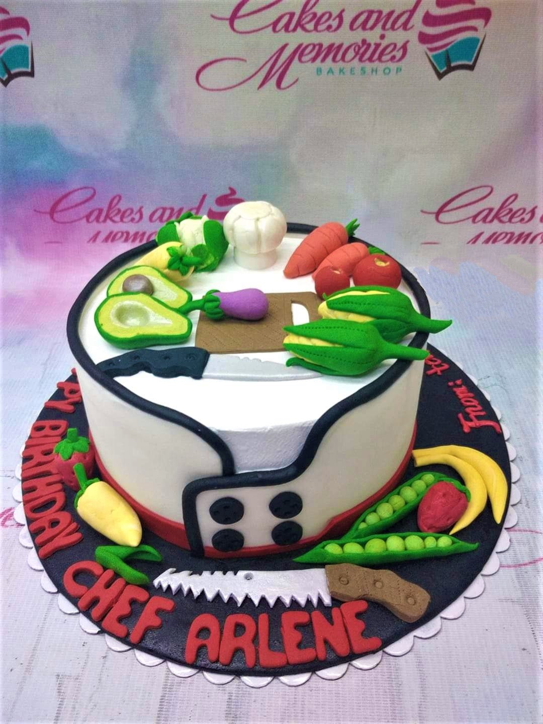 Chef-themed birthday cake for Chef Arlene featuring fondant vegetables, a chef's hat, and a knife on a white fondant base with black trim. Chef-themed birthday cake for Chef Arlene featuring fondant vegetables, a chef's hat, and a knife on a white fondant base with black trim.