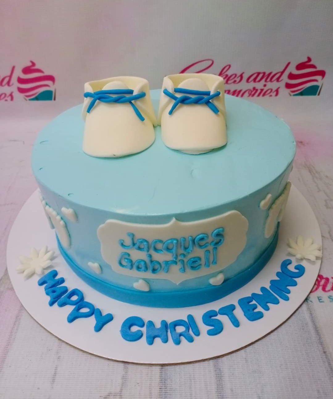 Light blue christening cake with white fondant baby shoes and blue laces on top, featuring a name plaque and message on the base board. Light blue christening cake with white fondant baby shoes and blue laces on top, featuring a name plaque and message on the base board.