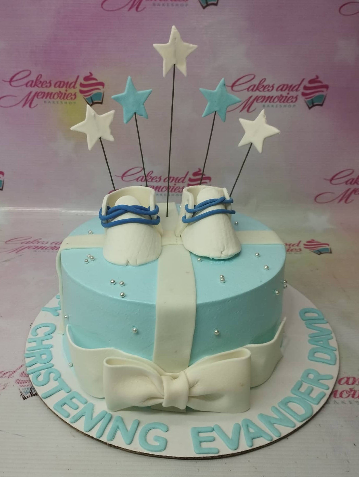 Light blue christening cake for Evander David featuring white fondant baby shoes and star toppers with a large white bow. Light blue christening cake for Evander David featuring white fondant baby shoes and star toppers with a large white bow.