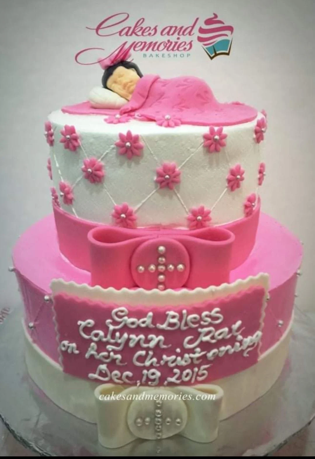 Christening Floral 2-Tier Cake Two-tier pink and white christening cake with a sleeping baby topper, pink fondant flowers, and large bows.