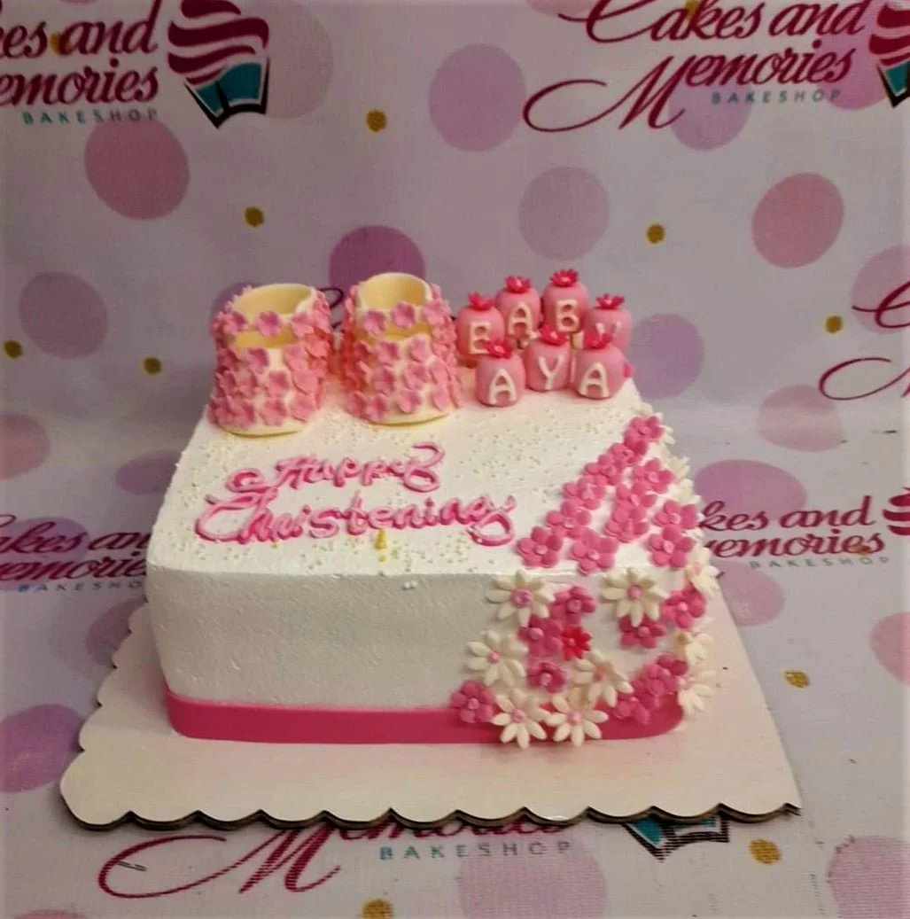 Christening White Floral Cake Square white christening cake with pink fondant baby shoes, pink flower decorations, and a pink message.