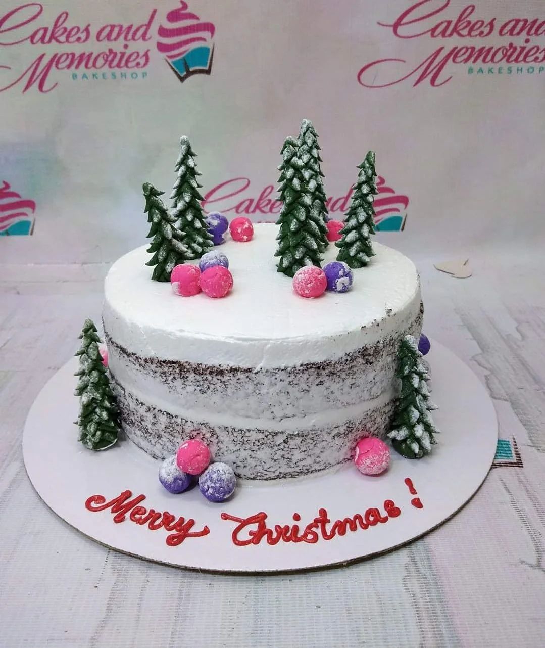 Semi-naked chocolate cake with white icing, green fondant pine trees, and pink and purple fondant balls for Christmas.