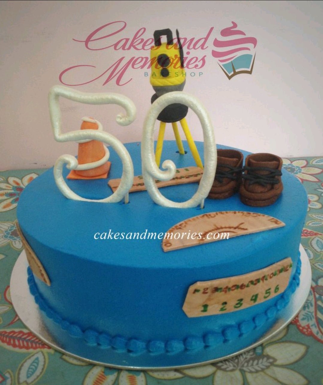 Blue 1-tier civil engineer themed birthday cake with 3D fondant surveying equipment, safety cone, boots, and protractor decorations. Blue 1-tier civil engineer themed birthday cake with 3D fondant surveying equipment, safety cone, boots, and protractor decorations.