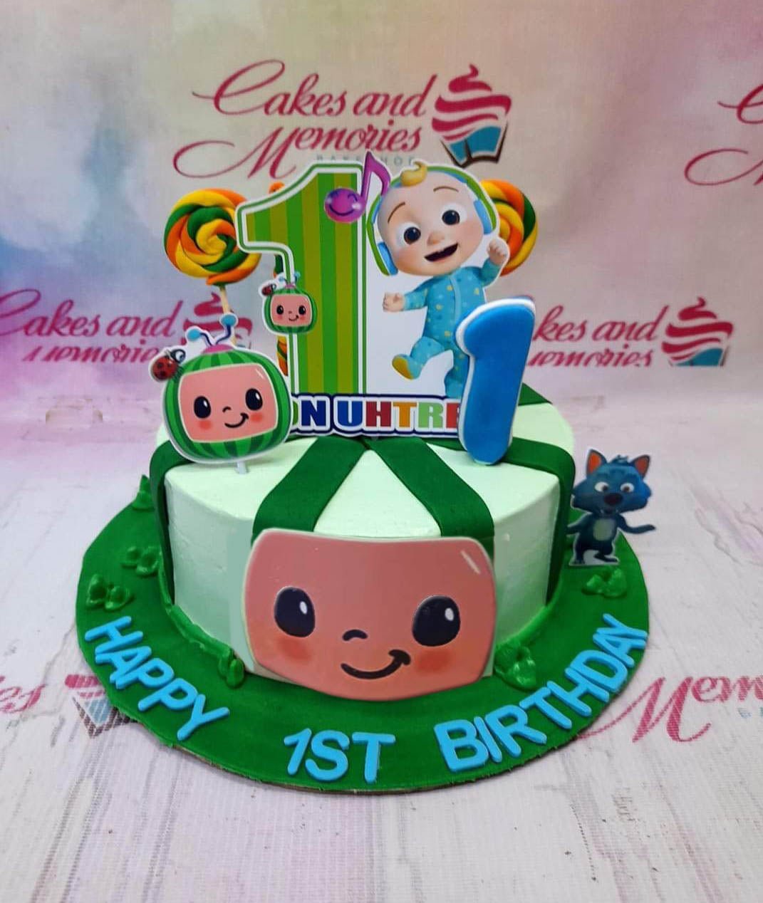 Cocomelon themed 1-tier birthday cake with green stripes, character printouts, and a blue number one candle on a green fondant base board. Cocomelon themed 1-tier birthday cake with green stripes, character printouts, and a blue number one candle on a green fondant base board.