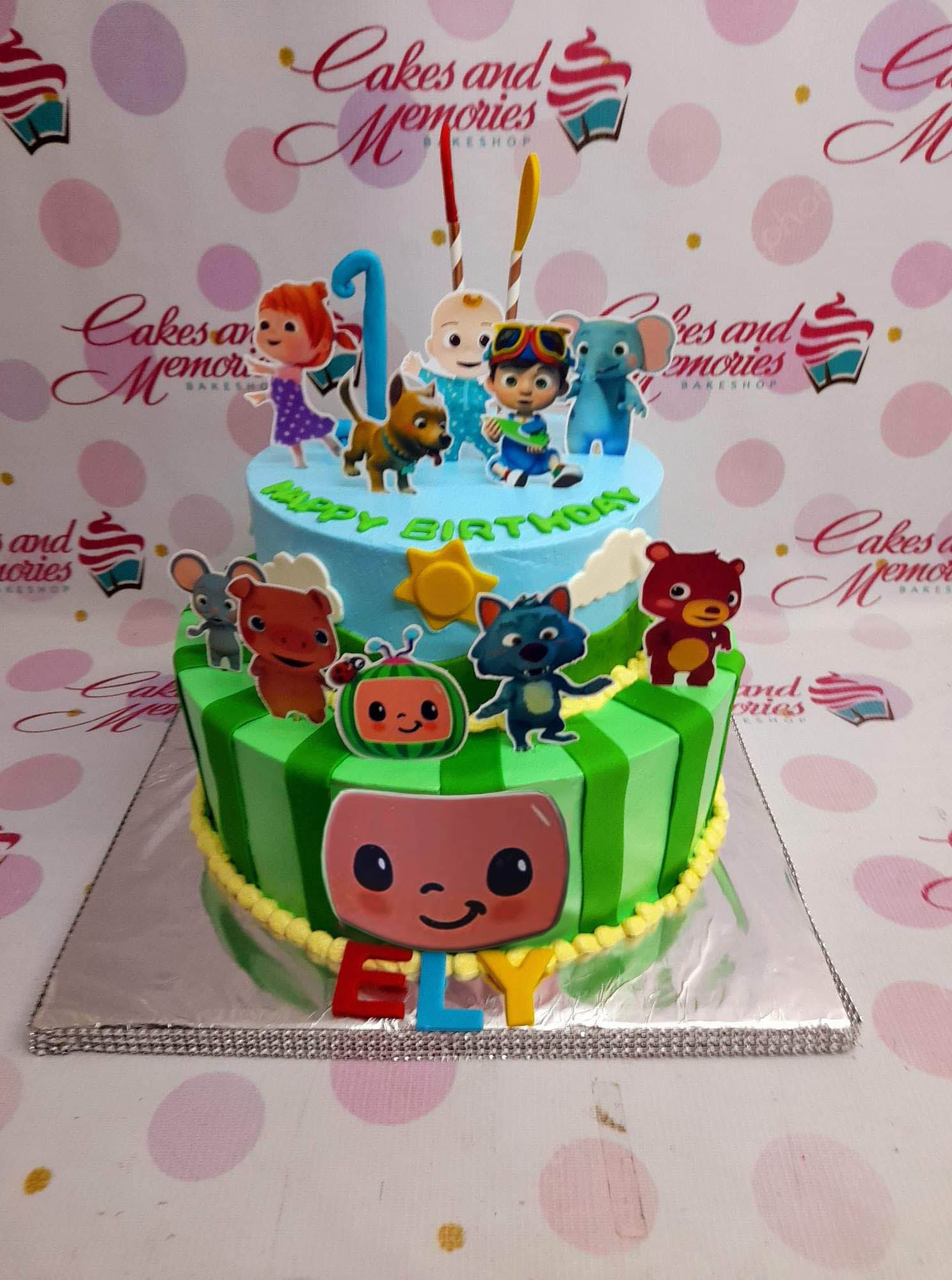 Two-tier Cocomelon themed birthday cake with blue and green striped icing, character printouts, and a yellow sun topper. Two-tier Cocomelon themed birthday cake with blue and green striped icing, character printouts, and a yellow sun topper.