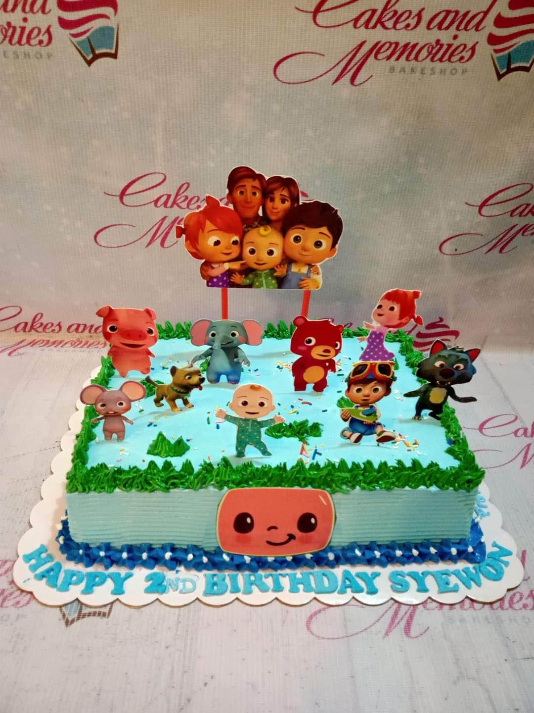 Cocomelon themed rectangle cake with light blue icing, green grass borders, and various character printouts including JJ and family. Cocomelon themed rectangle cake with light blue icing, green grass borders, and various character printouts including JJ and family.