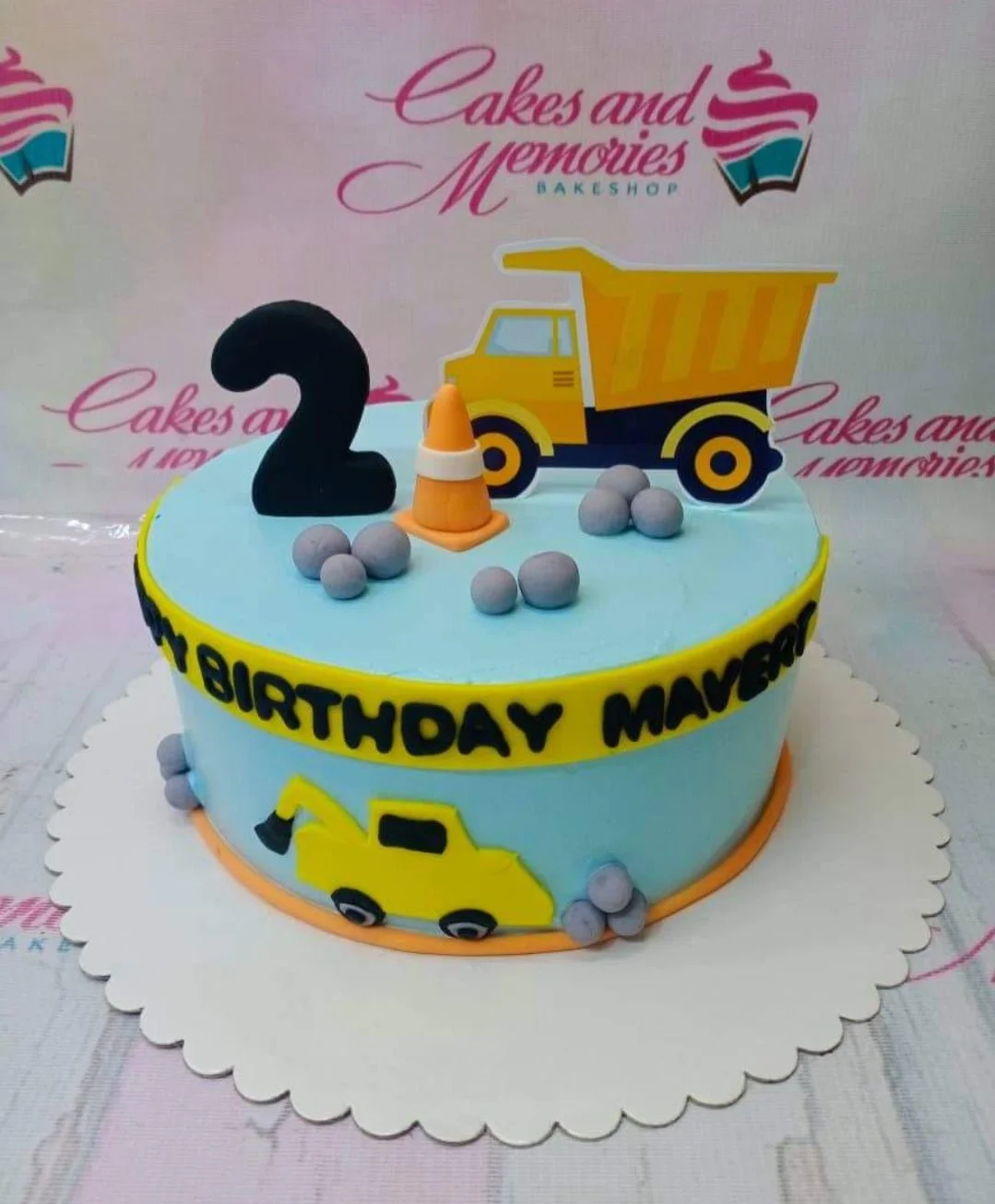 Construction Sky Blue Birthday Cake Light blue construction themed birthday cake with a yellow dump truck printout, orange traffic cone, and grey fondant rocks.