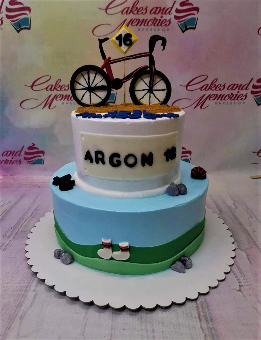 Two-tier cycling themed cake with a blue bottom tier and white top tier featuring a bicycle topper and Argon 18 branding.