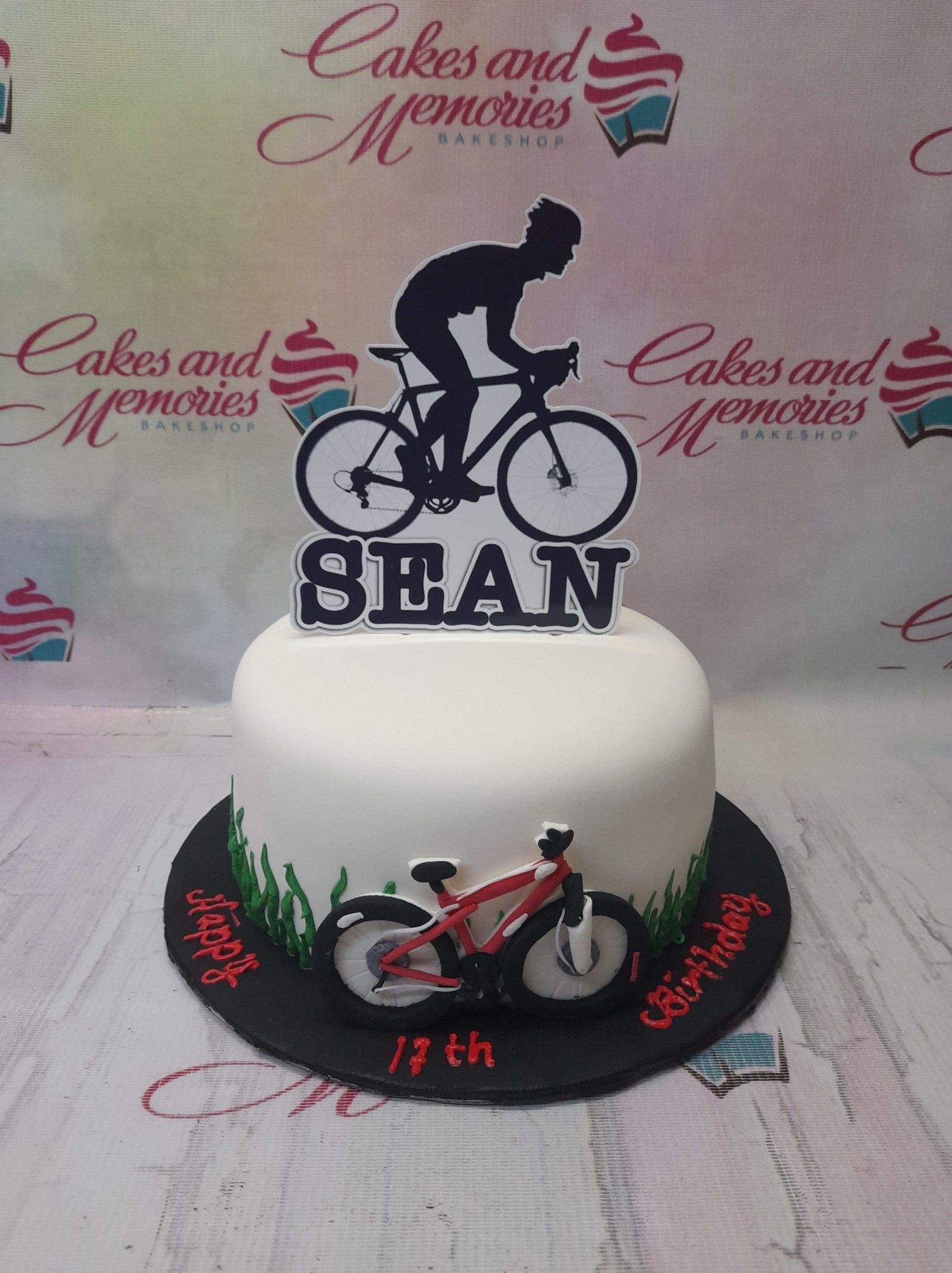 White fondant cyclist birthday cake with a black bicycle silhouette topper and a red 3D fondant mountain bike on the base board. White fondant cyclist birthday cake with a black bicycle silhouette topper and a red 3D fondant mountain bike on the base board.