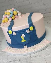 Pink denim overall themed first birthday cake with blue fondant overalls, yellow buttons, and a cluster of colorful balloon spheres. Pink denim overall themed first birthday cake with blue fondant overalls, yellow buttons, and a cluster of colorful balloon spheres.