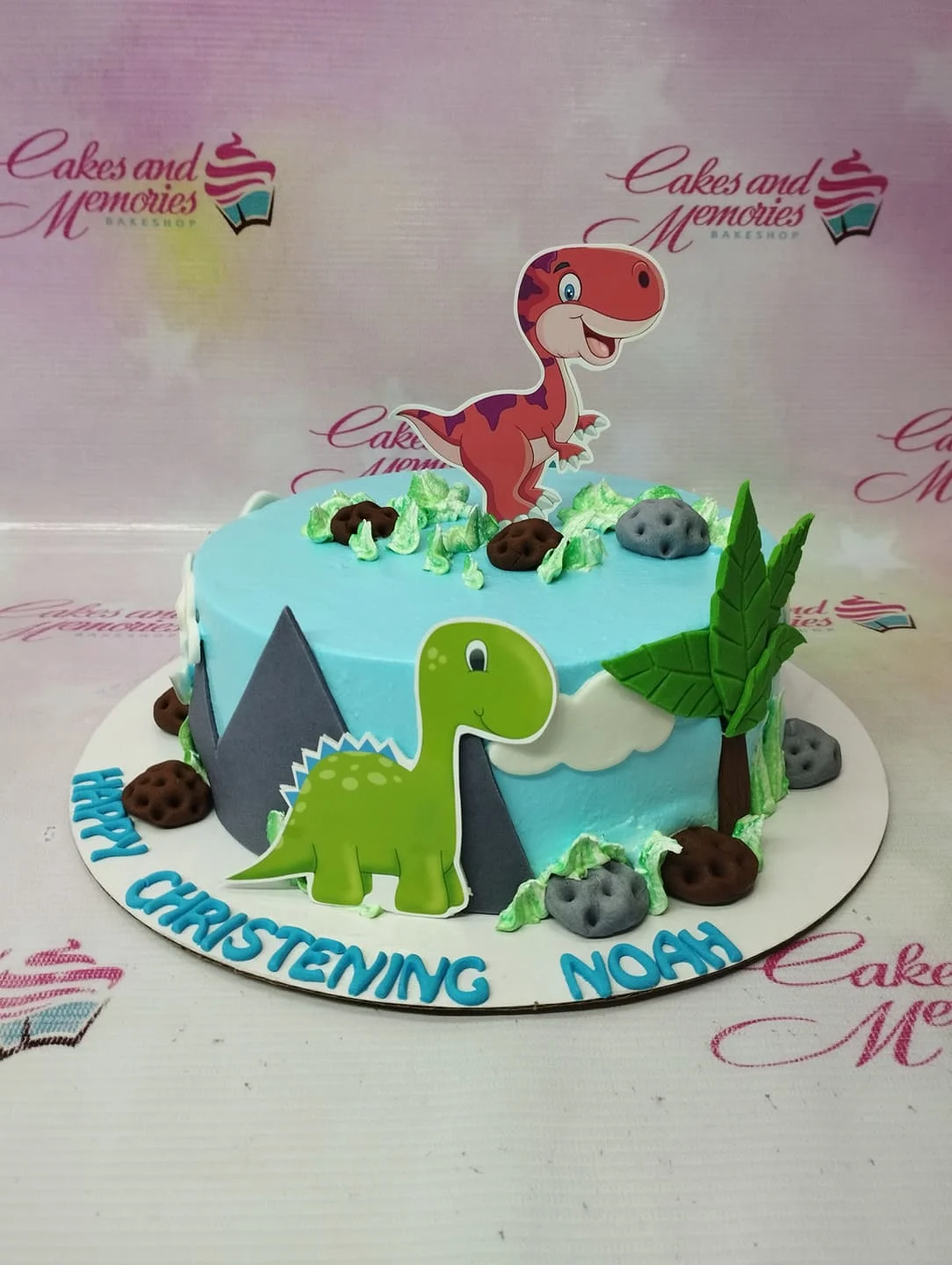 Light blue dinosaur themed christening cake with a red T-Rex topper, green dinosaur cutout, and fondant mountains and trees.