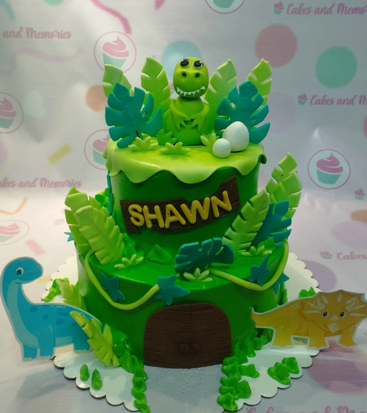 Two-tier green dinosaur themed cake with a fondant T-Rex topper, jungle leaves, and printed dinosaur cutouts.