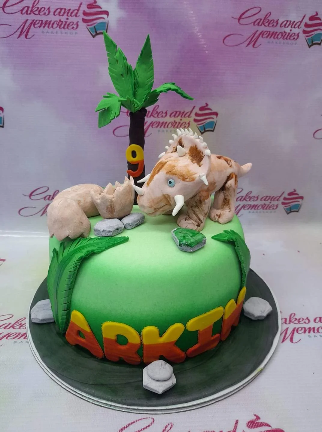 Green fondant dinosaur cake with a 3D triceratops figure, palm tree, and gumpaste rocks for a 9th birthday celebration.