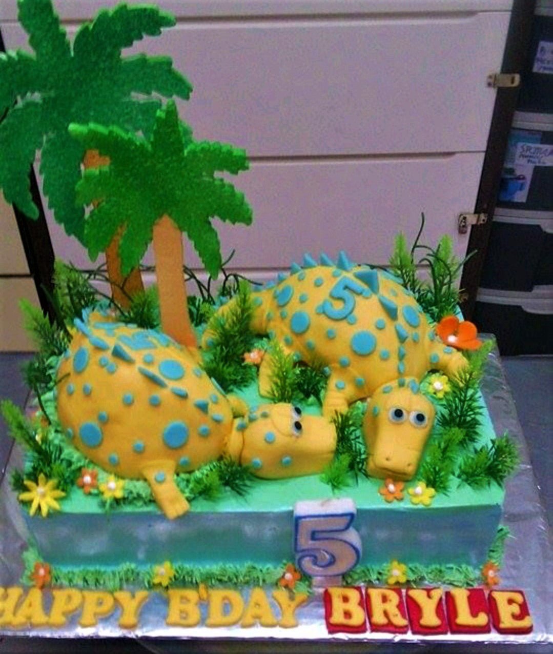 Dinosaur themed rectangle cake with yellow 3D fondant dinosaurs, palm trees, and green icing grass for a 5th birthday. Dinosaur themed rectangle cake with yellow 3D fondant dinosaurs, palm trees, and green icing grass for a 5th birthday.