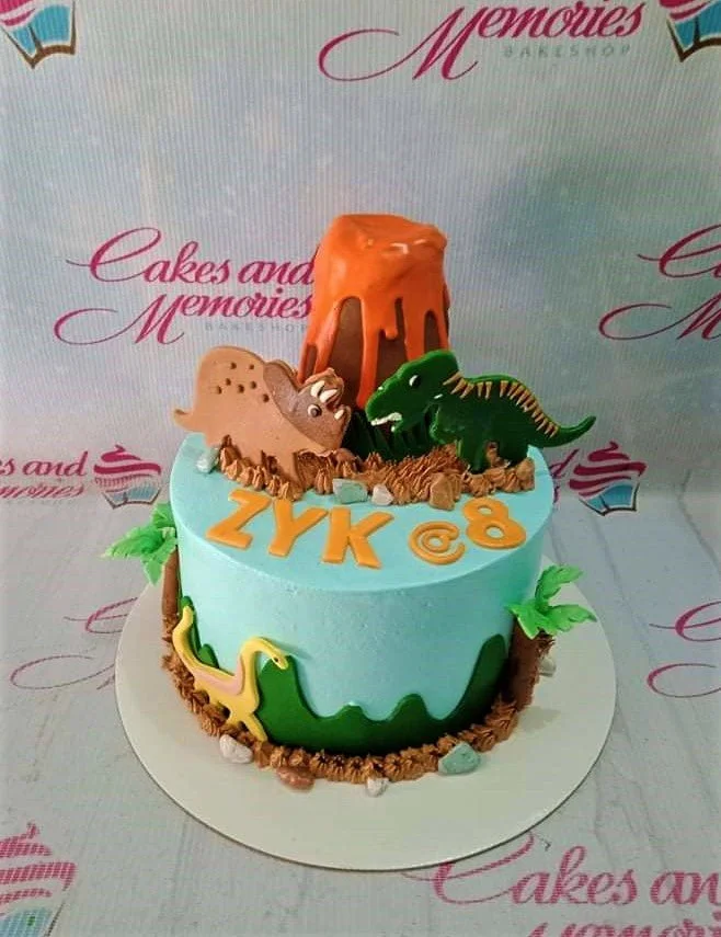 Light blue dinosaur themed birthday cake with a fondant volcano, T-Rex, and Triceratops toppers.