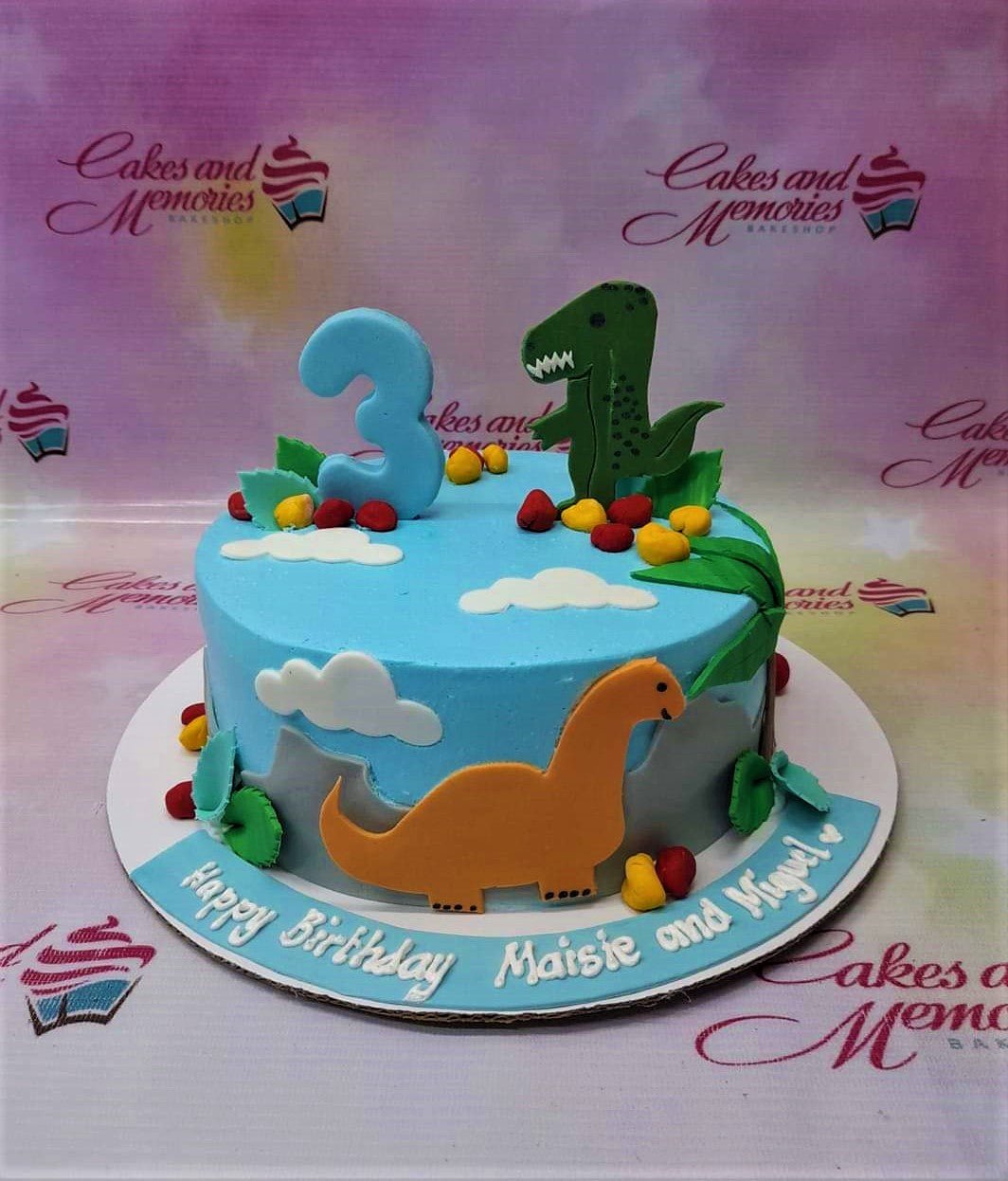 Light blue dinosaur themed birthday cake with a green T-Rex, orange Brontosaurus, and a blue number 3 topper. Light blue dinosaur themed birthday cake with a green T-Rex, orange Brontosaurus, and a blue number 3 topper.