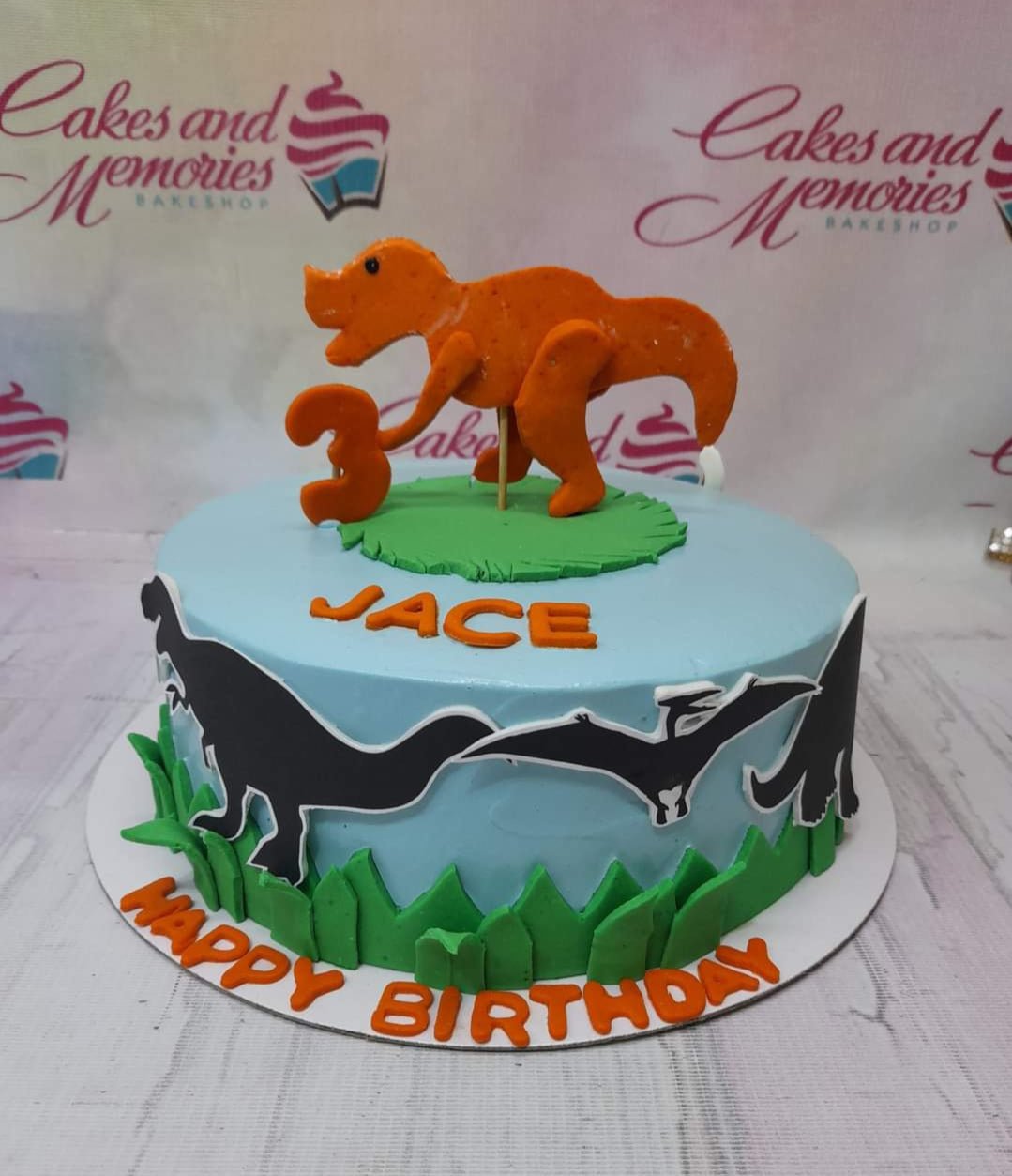 Light blue dinosaur birthday cake with an orange fondant T-Rex topper, green grass accents, and black dinosaur silhouettes on the side. Light blue dinosaur birthday cake with an orange fondant T-Rex topper, green grass accents, and black dinosaur silhouettes on the side.