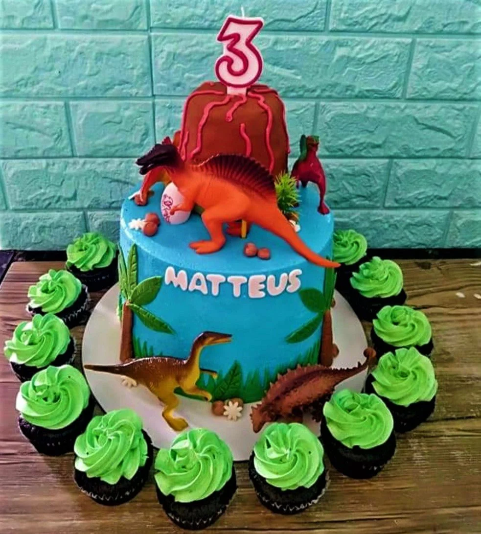 Two-tier dinosaur themed birthday cake with a brown volcano top tier, blue bottom tier, and plastic dinosaur toys.