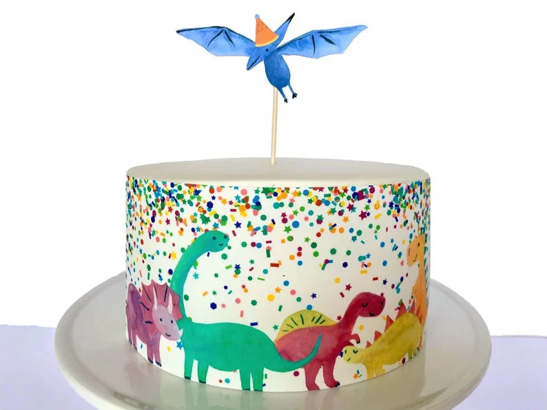 White fondant dinosaur birthday cake featuring colorful printed dinosaur cutouts and a blue pterodactyl topper with confetti patterns.