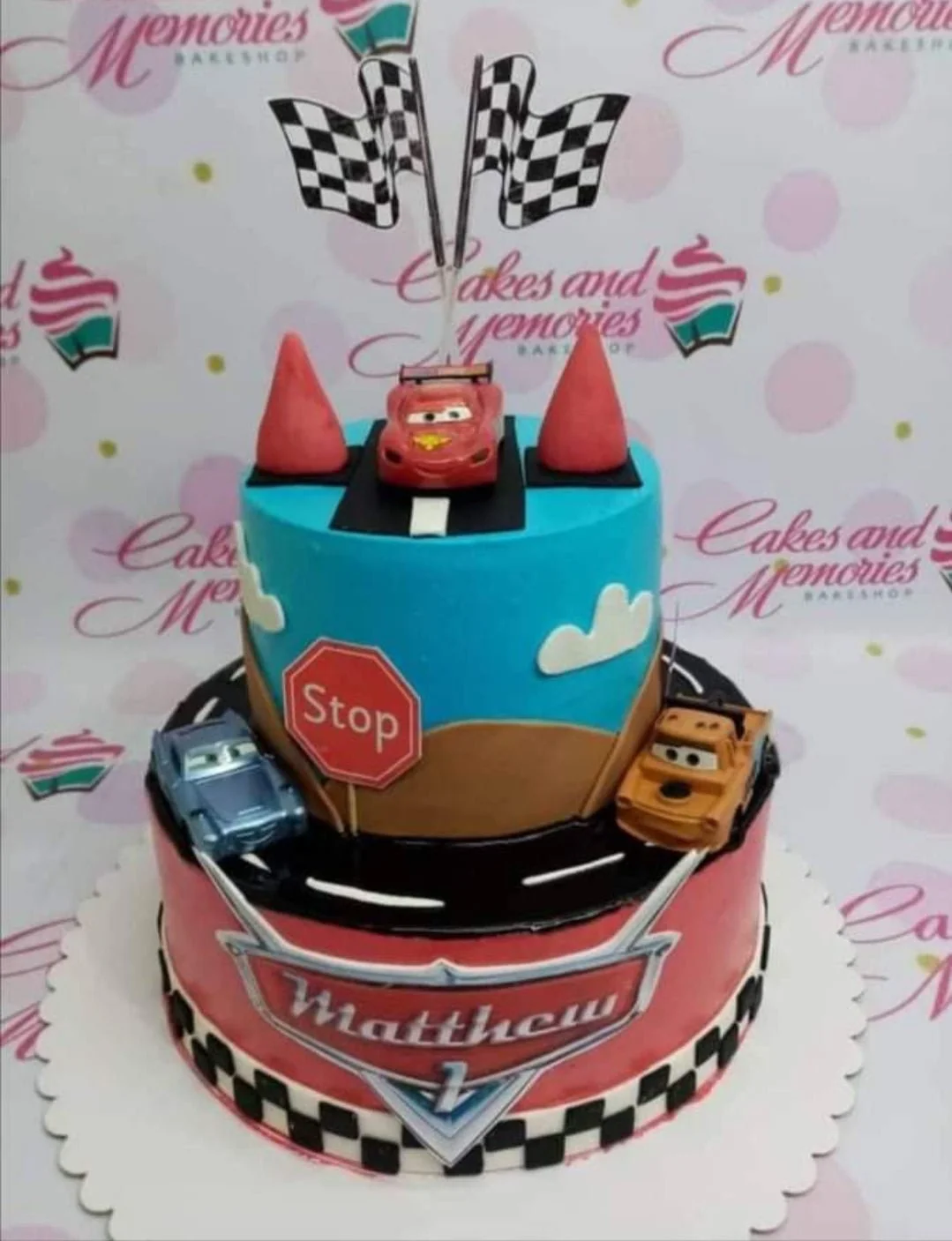Disney-Inspired Cars 2-Tier Birthday Cake Two-tier Disney Cars themed birthday cake with Lightning McQueen, Mater, and Sally figurines on a racing track design.