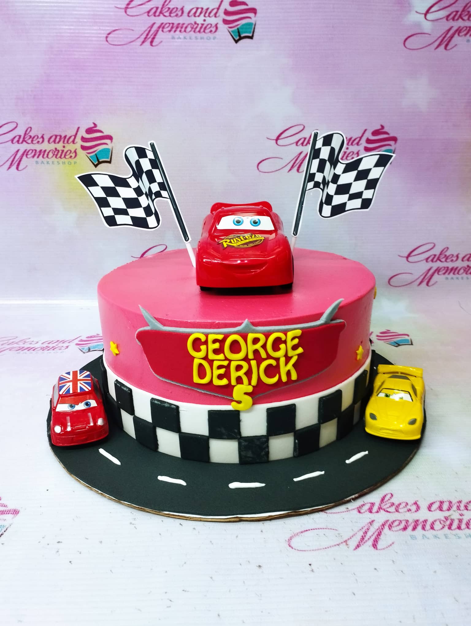 Red Disney Cars themed birthday cake with Lightning McQueen toy and checkered flag printouts on a black road base board. Red Disney Cars themed birthday cake with Lightning McQueen toy and checkered flag printouts on a black road base board.
