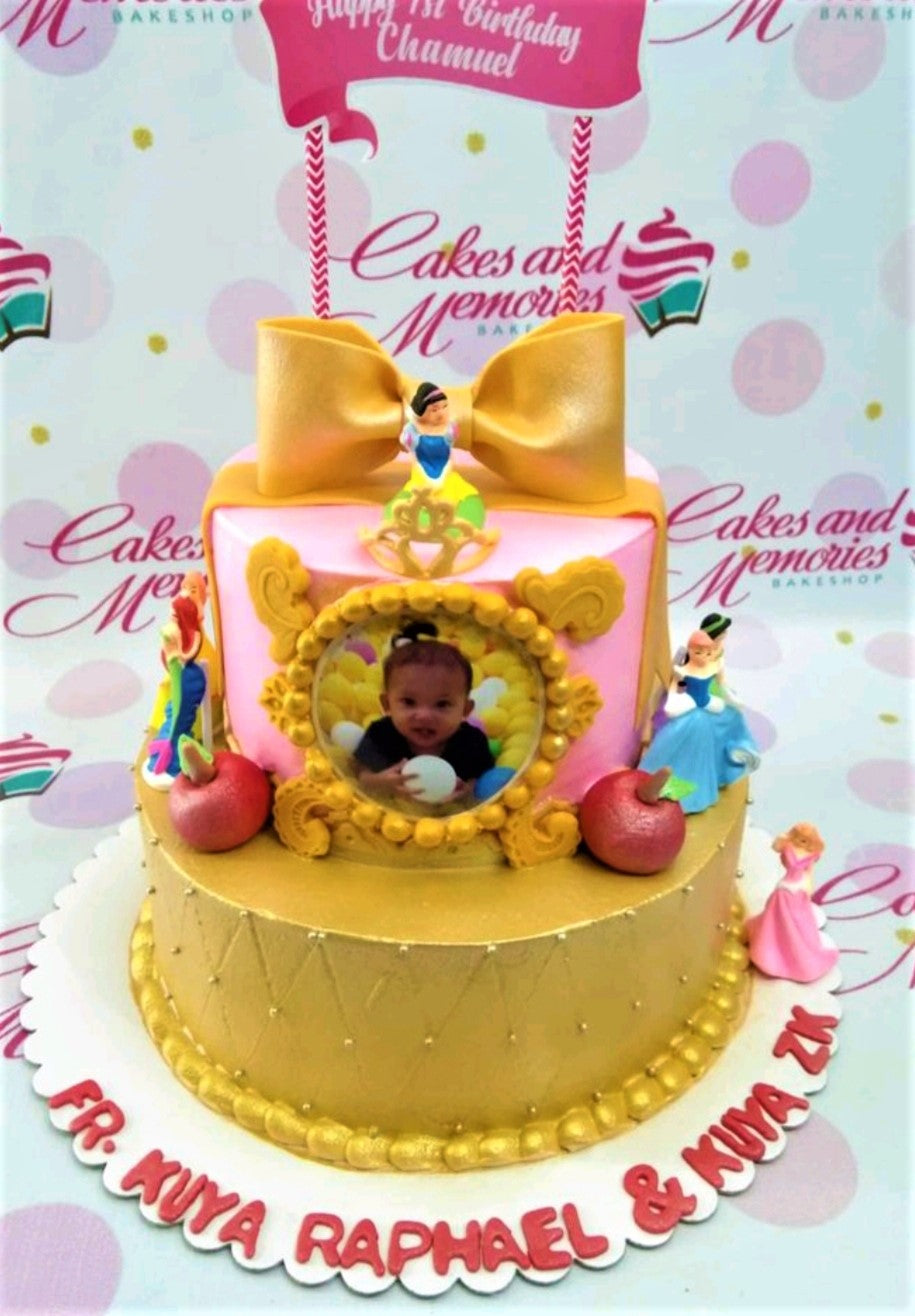 Two-tier Disney Princess themed cake with a pink top tier, gold bottom tier, a large gold bow, and various princess figurines. Two-tier Disney Princess themed cake with a pink top tier, gold bottom tier, a large gold bow, and various princess figurines.