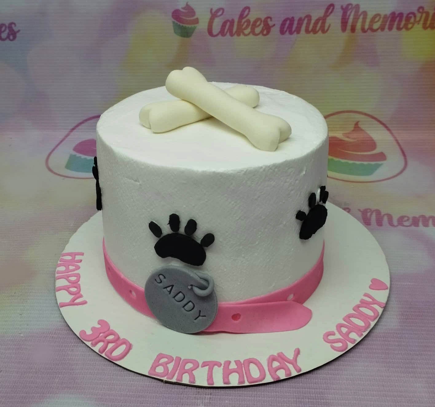 White soft-icing dog-themed cake with fondant bones on top, black paw prints on the side, and a pink fondant collar with a silver name tag. White soft-icing dog-themed cake with fondant bones on top, black paw prints on the side, and a pink fondant collar with a silver name tag.