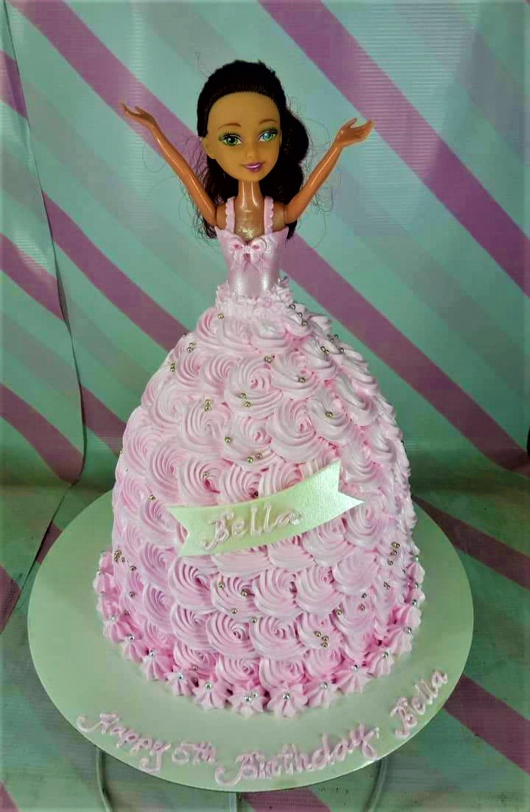 Pink doll cake with a doll topper and pink icing rosettes forming a dress, decorated with silver dragees and a name banner. Pink doll cake with a doll topper and pink icing rosettes forming a dress, decorated with silver dragees and a name banner.