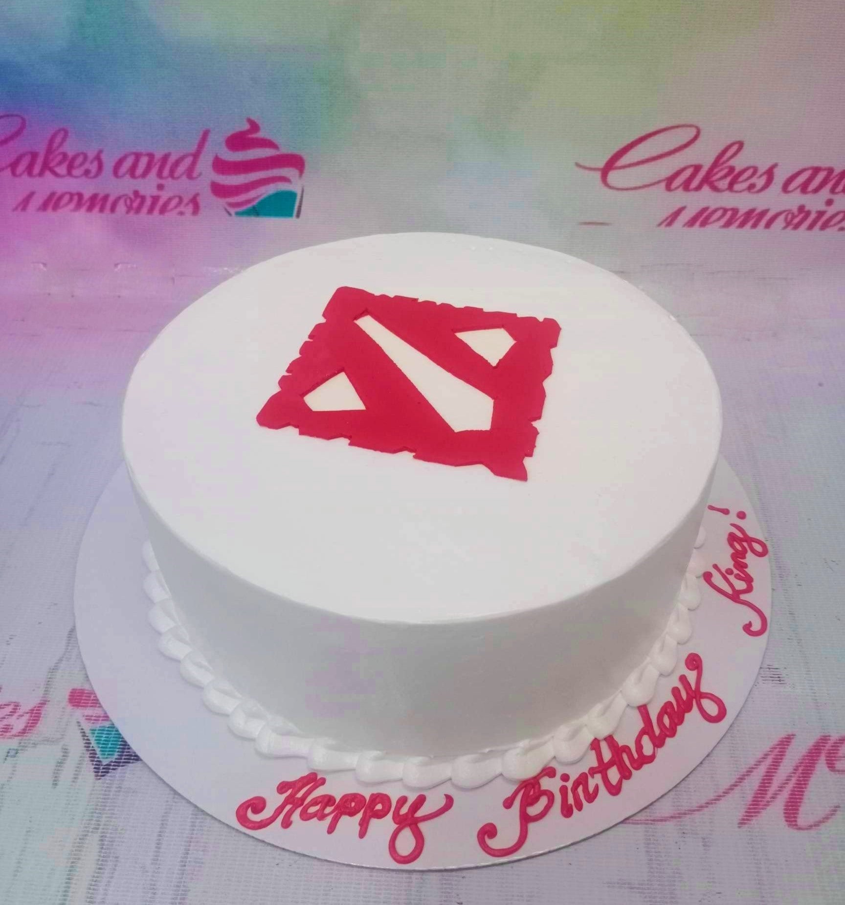 White minimalist Dota 2 themed birthday cake with a red logo on top and pink icing message on the base board. White minimalist Dota 2 themed birthday cake with a red logo on top and pink icing message on the base board.