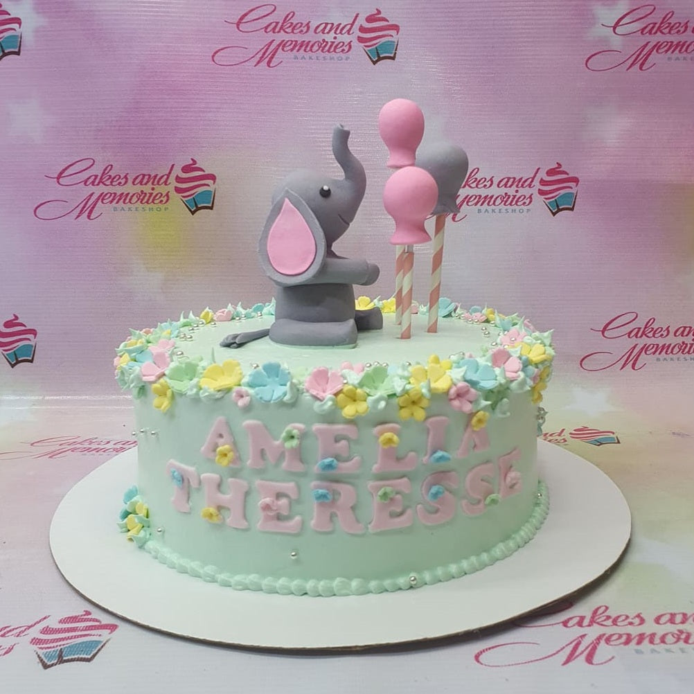 Light green birthday cake with a 3D fondant elephant topper, pink balloons, and a floral icing border with the name Amelia Therese. Light green birthday cake with a 3D fondant elephant topper, pink balloons, and a floral icing border with the name Amelia Therese.