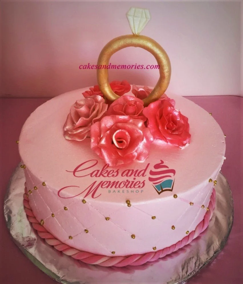 Engagement Pink Floral Cake Pink round cake with a large gold ring topper, pink fondant roses, and a quilted pattern with gold dragees on the side.