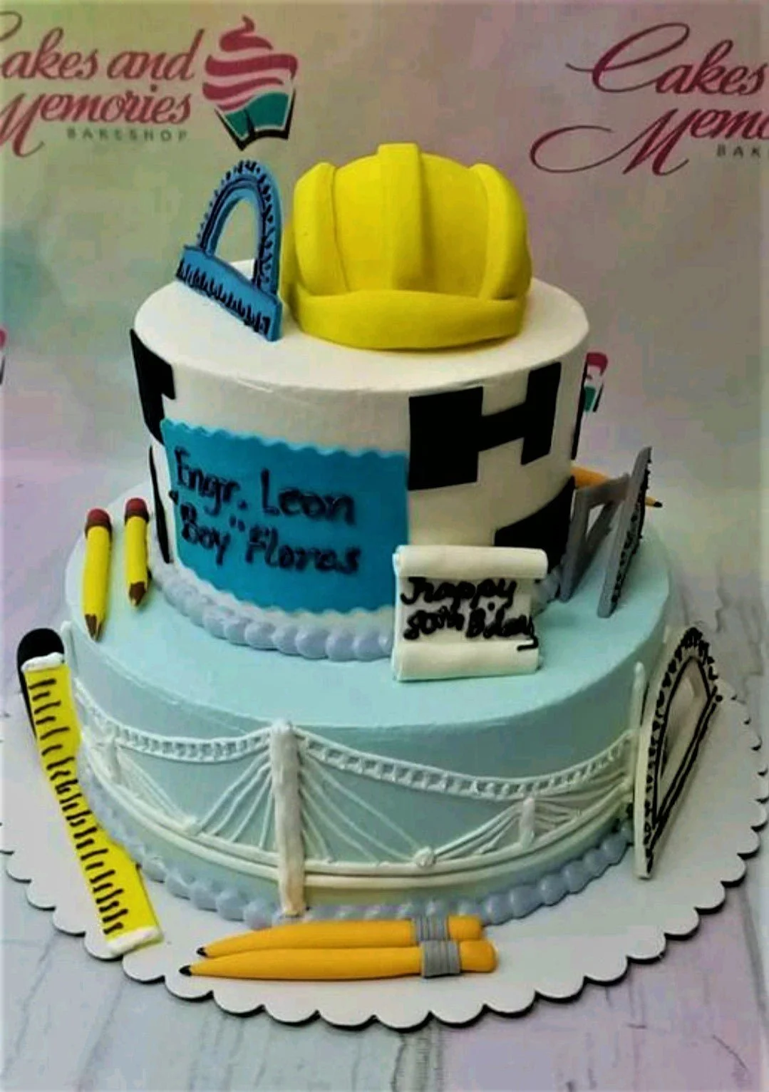 2-tier blue and white engineering themed cake with a yellow hard hat, pencils, and bridge design.