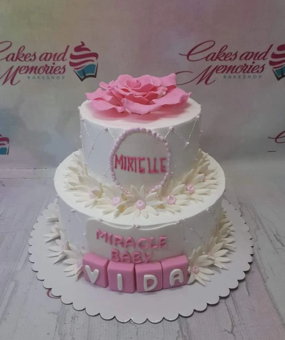 Floral Baby White 2-Tier Christening Cake White 2-tier floral cake with pink fondant rose, daisy accents, and pink block letters for a baby celebration.