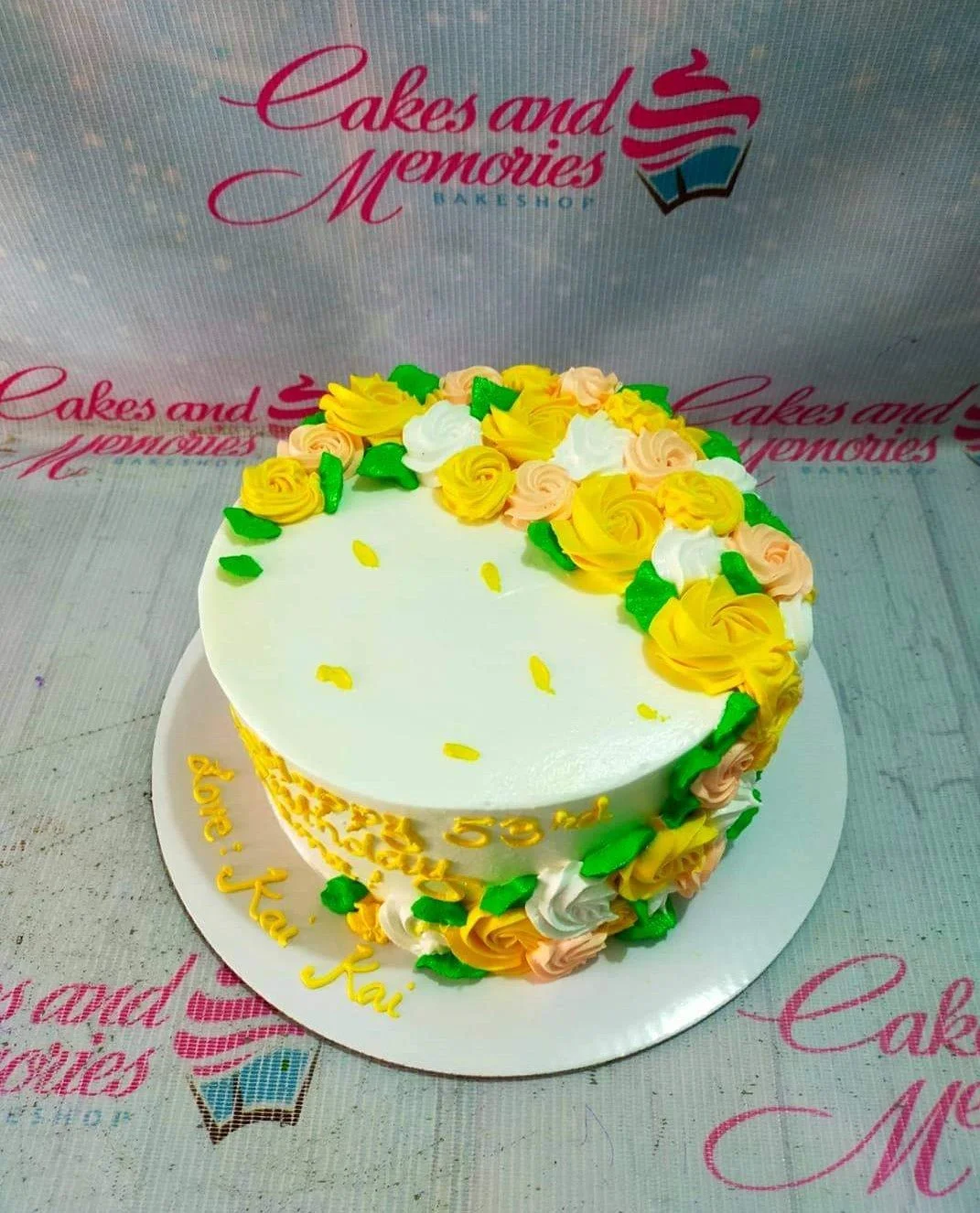 Floral Birthday White Cake A white 1 Tier round cake, Floral Birthday themed, featuring yellow, peach, and white icing roses, green leaves, and birthday text.