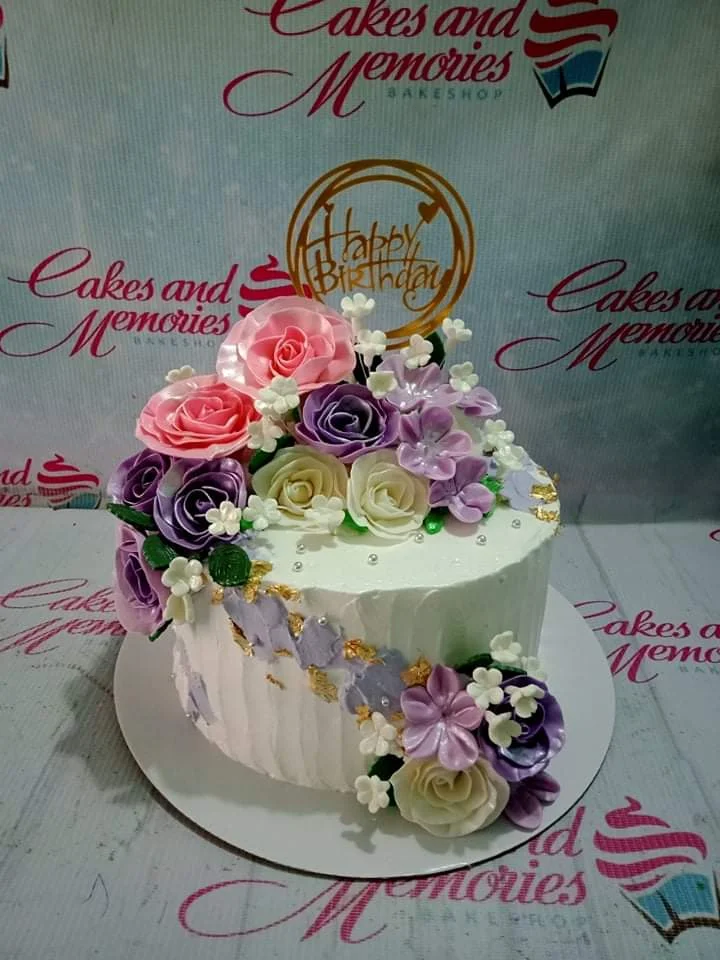 Floral Birthday White Debut Cake White soft-icing cake with vertical textured ridges, decorated with a lush bouquet of pink, purple, and white edible roses and gold leaf accents.