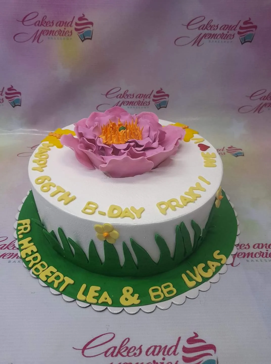Floral Birthday White Cake A white 1-tier cake themed around Floral Birthday, featuring a large pink gumpaste flower and green grass icing with yellow letters.