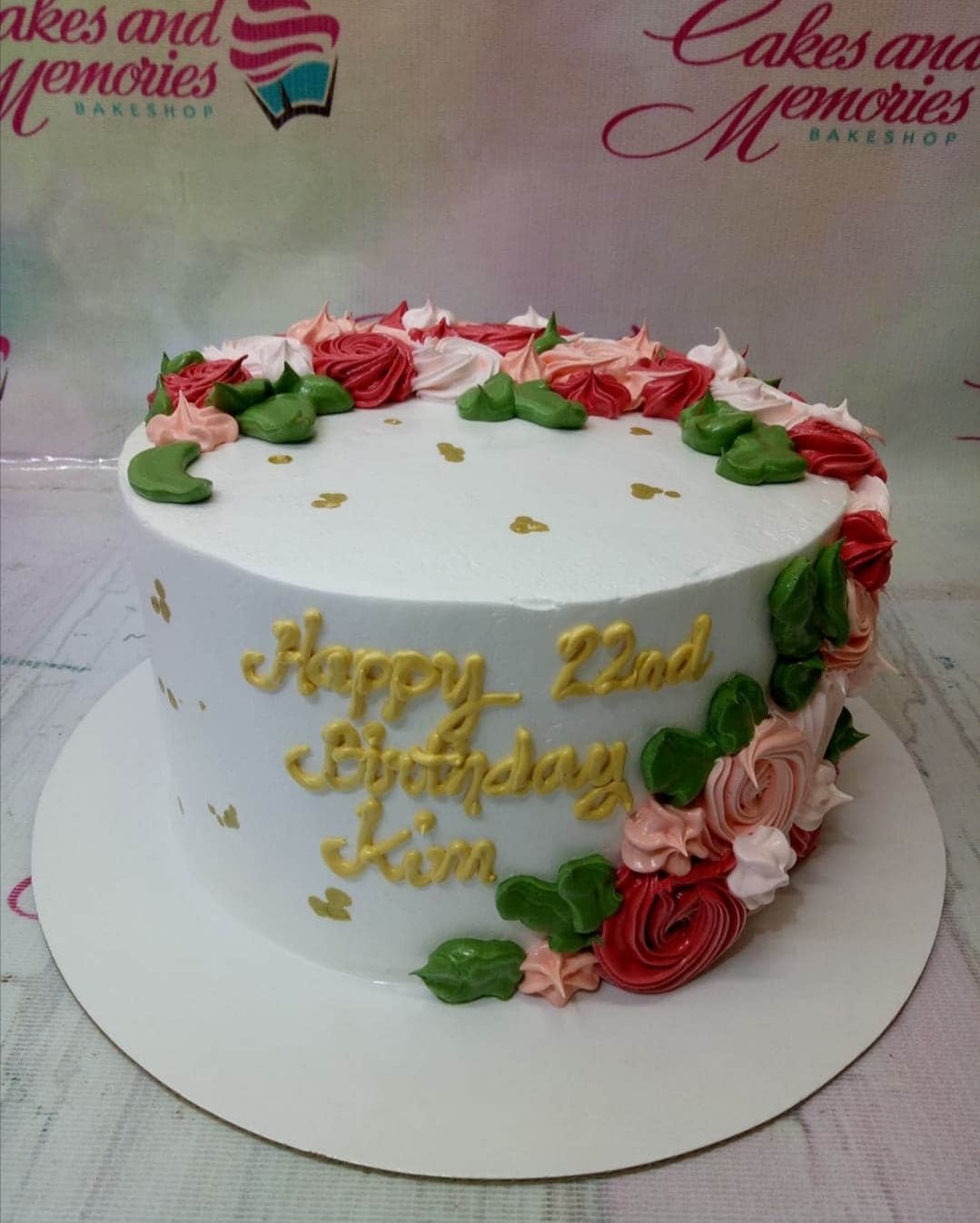 White round birthday cake with pink and red icing rosettes, green leaves, and gold lettering for Kim's 22nd birthday.