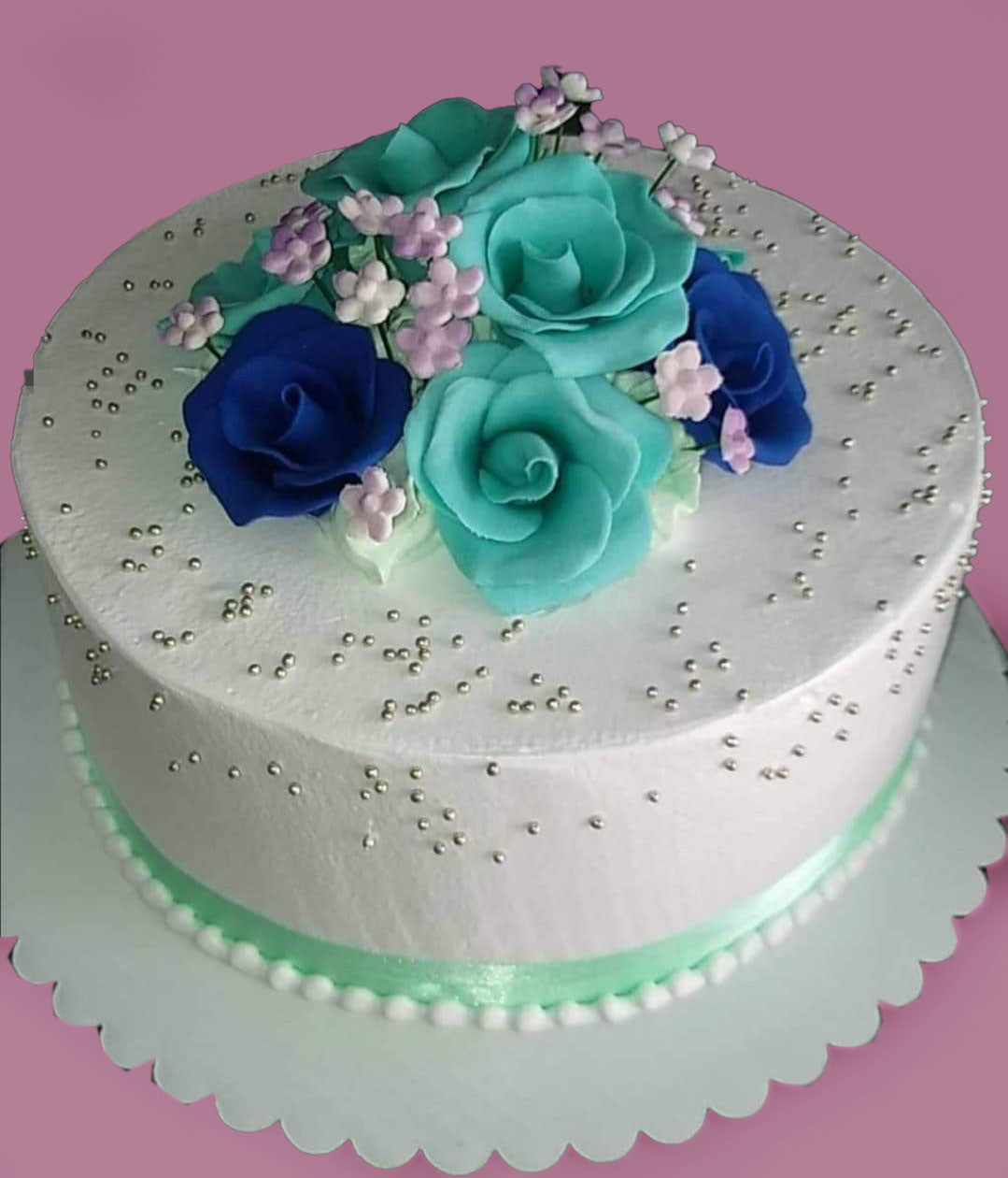 White round cake with teal and navy blue gumpaste roses, purple filler flowers, and silver dragees. White round cake with teal and navy blue gumpaste roses, purple filler flowers, and silver dragees.