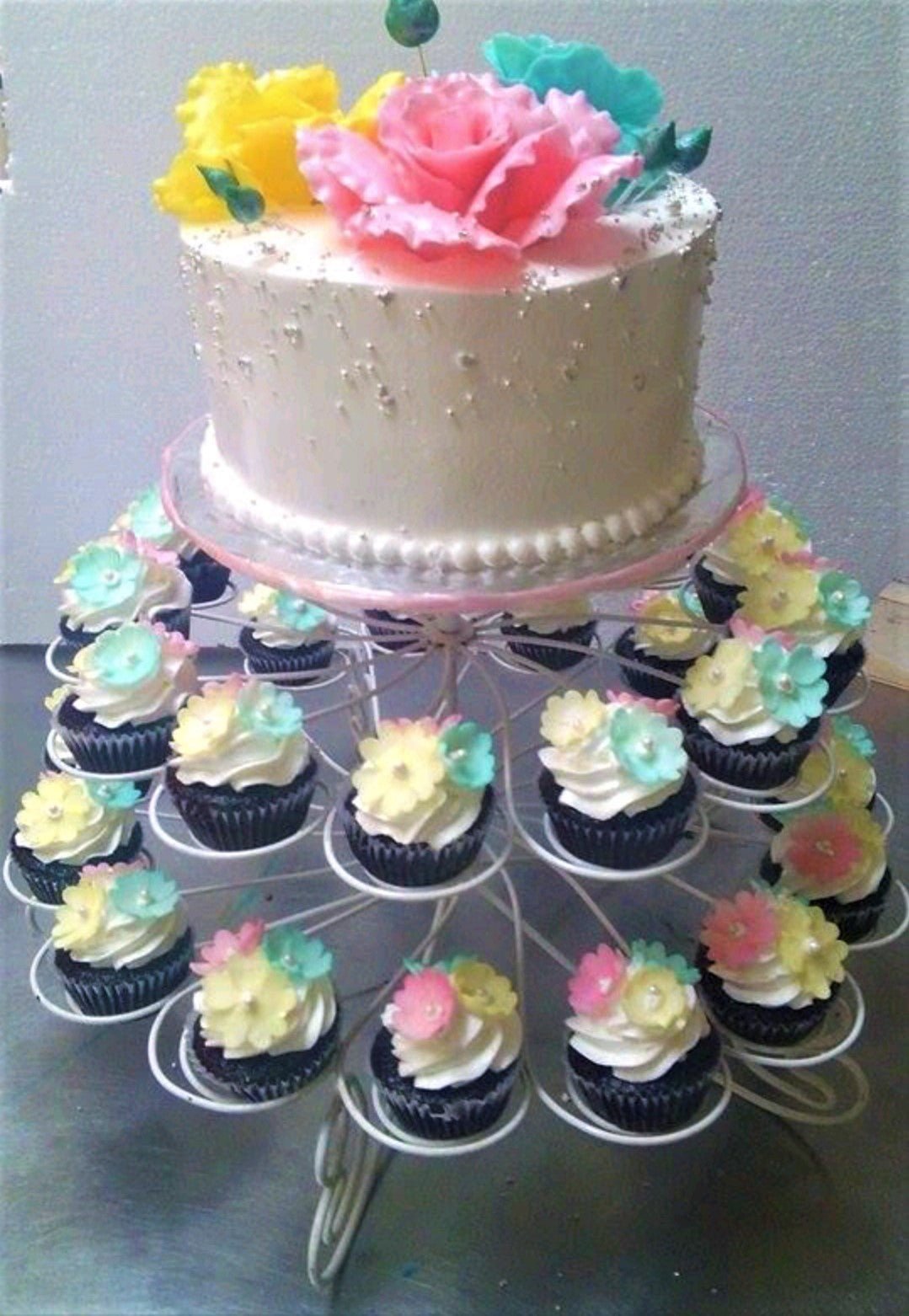 White 1-tier cake with large pink, yellow, and teal gumpaste roses on top, silver dragees on the sides, and matching floral cupcakes. White 1-tier cake with large pink, yellow, and teal gumpaste roses on top, silver dragees on the sides, and matching floral cupcakes.