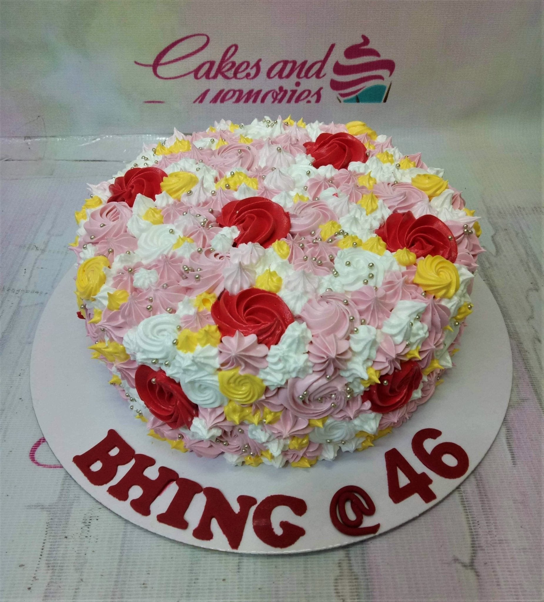 Round birthday cake covered in pink, white, yellow, and red icing rosettes with silver dragees and a red message on the base board. Round birthday cake covered in pink, white, yellow, and red icing rosettes with silver dragees and a red message on the base board.