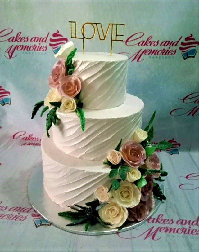Elegant three-tier white wedding cake with textured diagonal icing and a cascade of pink and cream edible roses topped with a gold LOVE sign. Elegant three-tier white wedding cake with textured diagonal icing and a cascade of pink and cream edible roses topped with a gold LOVE sign.