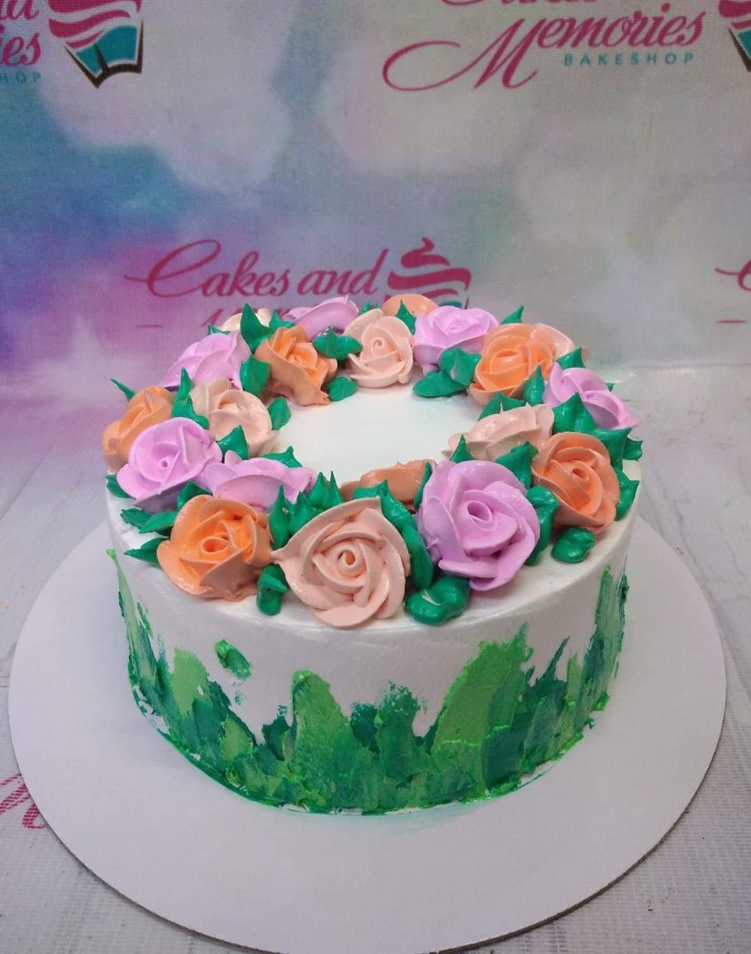 White round cake decorated with a wreath of pink, peach, and orange icing roses and green leaves with green palette knife textures on the side. White round cake decorated with a wreath of pink, peach, and orange icing roses and green leaves with green palette knife textures on the side.