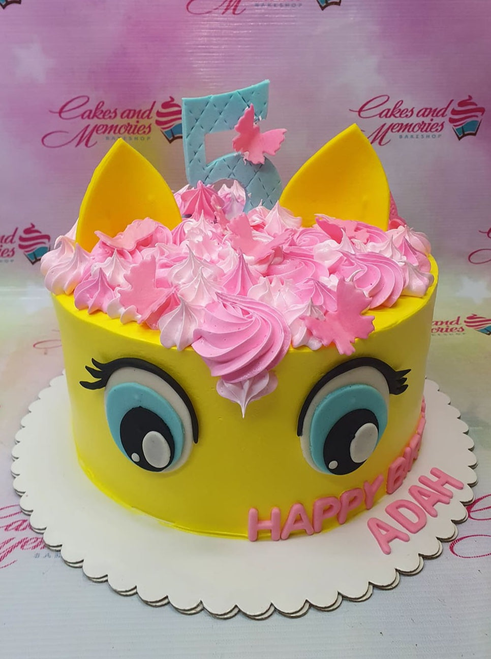 Yellow character-themed cake with pink and light pink icing swirls, large blue eyes, and a blue number 5 topper with a pink butterfly. Yellow character-themed cake with pink and light pink icing swirls, large blue eyes, and a blue number 5 topper with a pink butterfly.