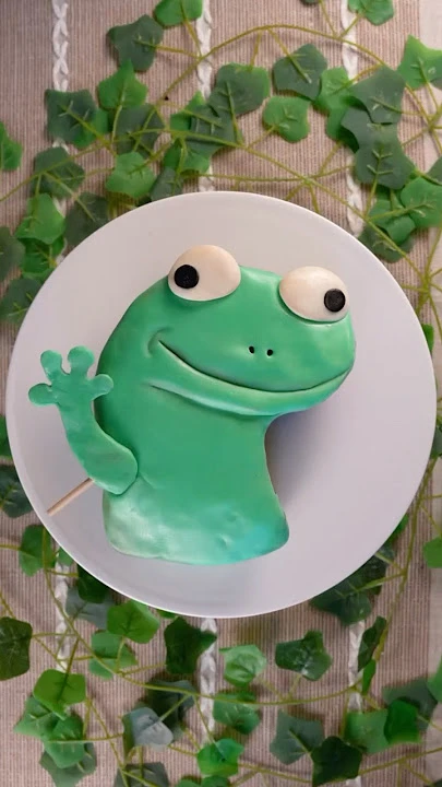 Green frog-shaped fondant cake with large white eyes and a waving hand on a white plate. Green frog-shaped fondant cake with large white eyes and a waving hand on a white plate.