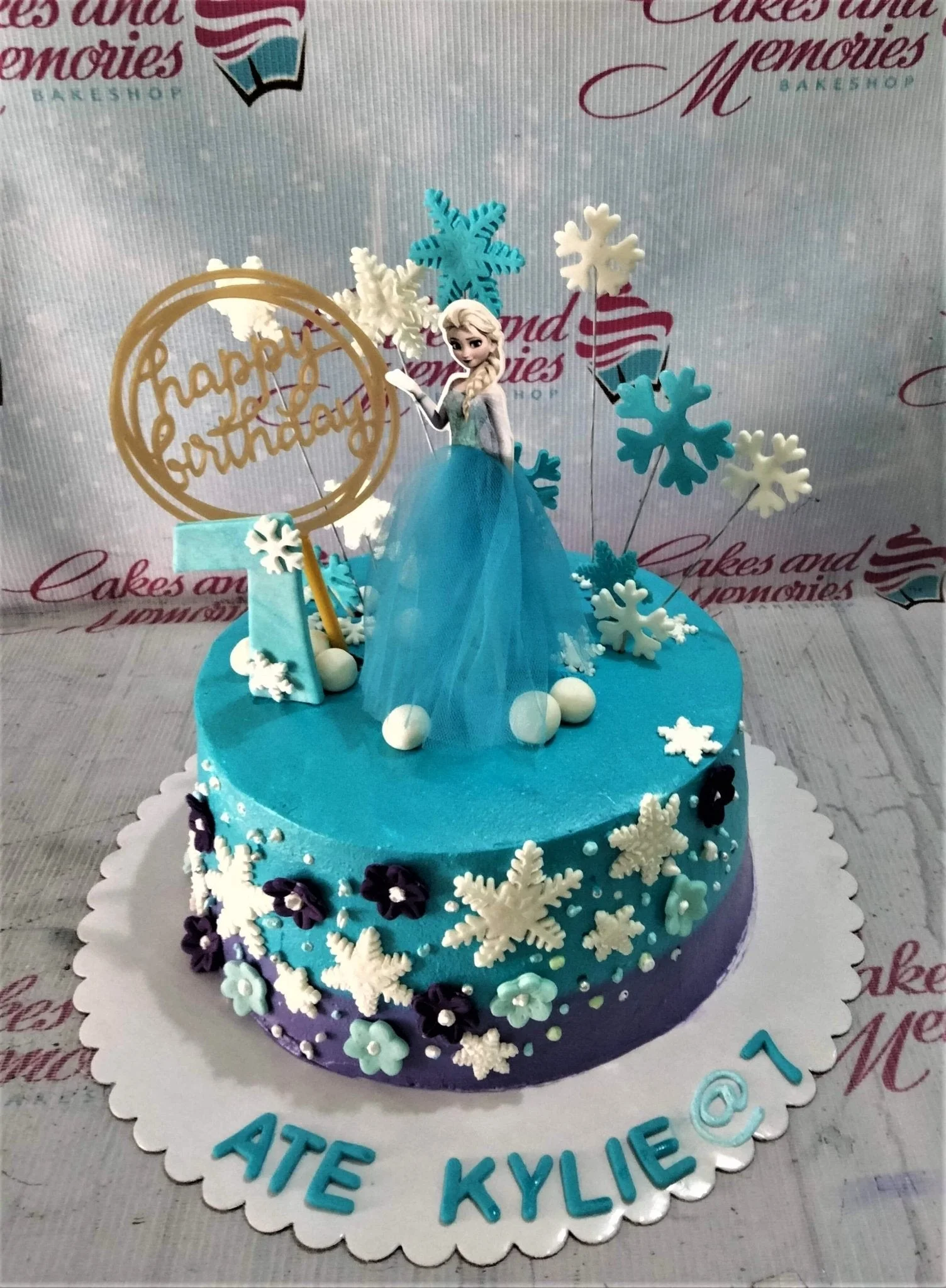 Frozen-Inspired Elsa Blue Birthday Cake A blue and purple 1-tier cake themed around Frozen Elsa, featuring an Elsa printout, gold Happy Birthday sign, and number 7 topper, with snowflakes and purple flowers.