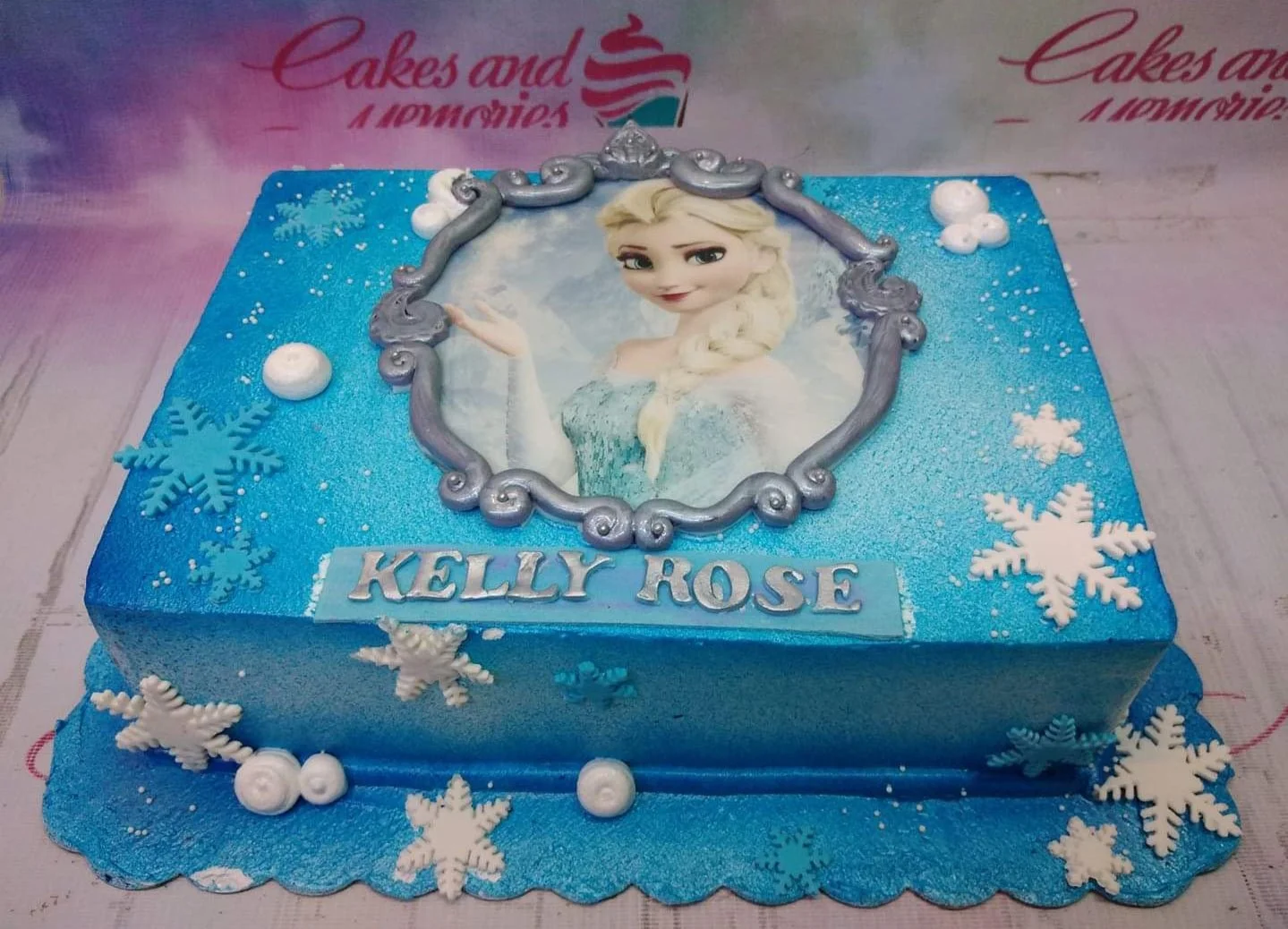 Frozen-Inspired Elsa Blue Floral Rectangle Cake A blue rectangle cake themed Frozen Elsa, featuring an Elsa portrait in a silver frame and white and blue snowflakes.