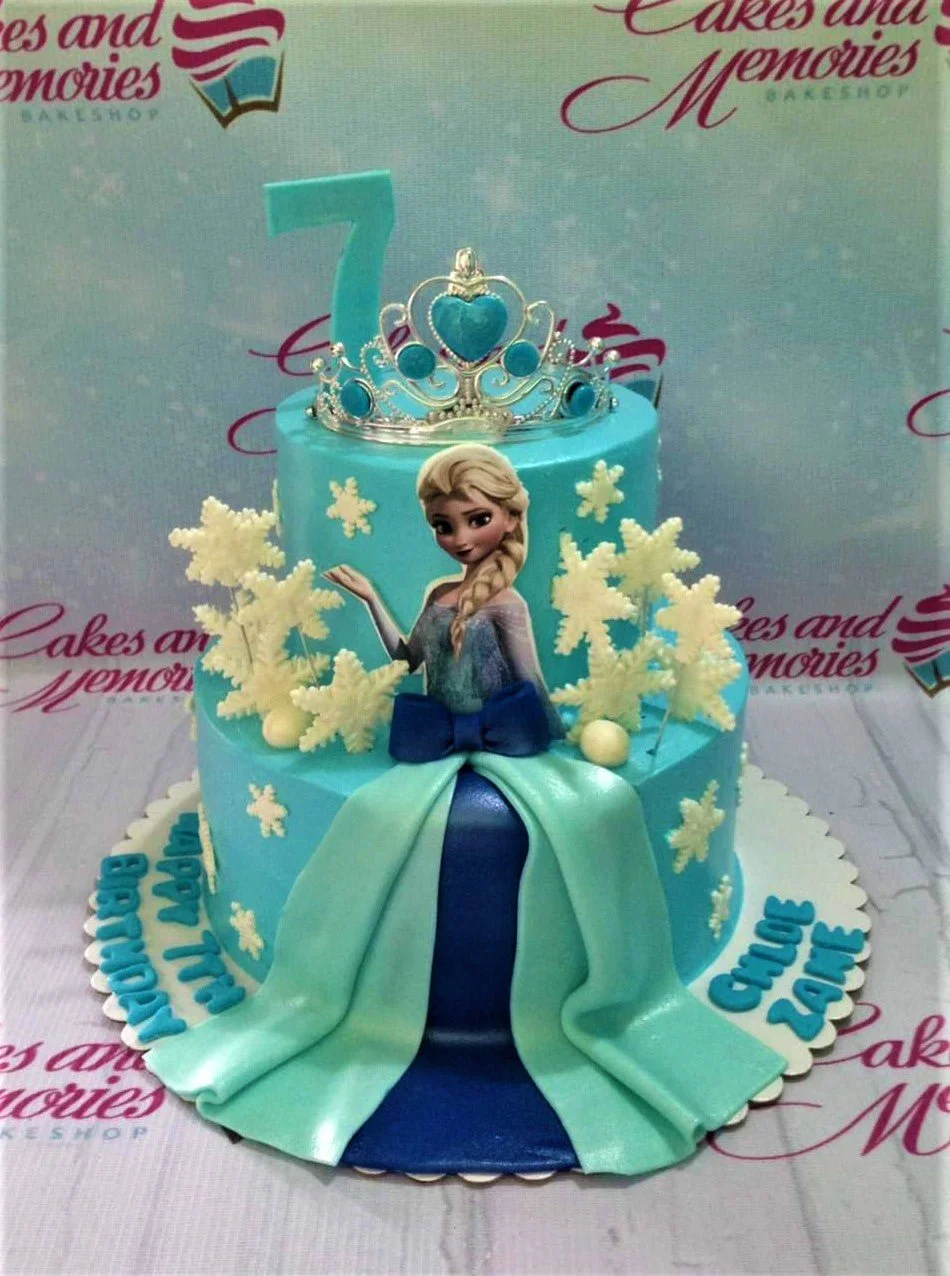 Two-tier light blue Frozen themed cake featuring Elsa printout, a silver rhinestone tiara, and white fondant snowflakes.