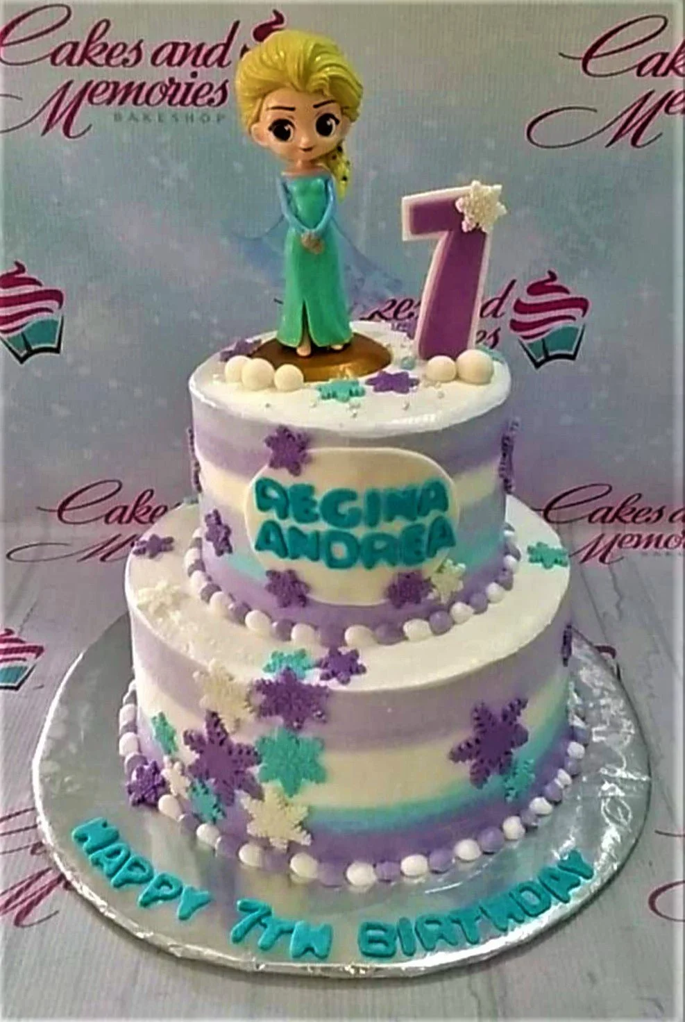 Frozen-Inspired Elsa 2-Tier Cake A white, purple, and blue two-tier cake themed around Frozen Elsa, featuring an Elsa figurine, a purple number seven, and various snowflake decorations.