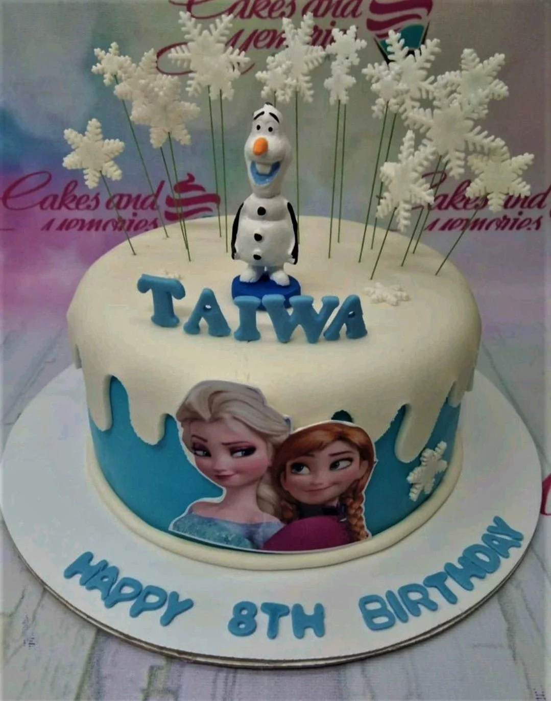Frozen-Inspired Birthday Cake A blue and white 1 Tier Fondant cake themed around Frozen, featuring an Olaf figurine, Elsa and Anna printouts, and white snowflake accents.