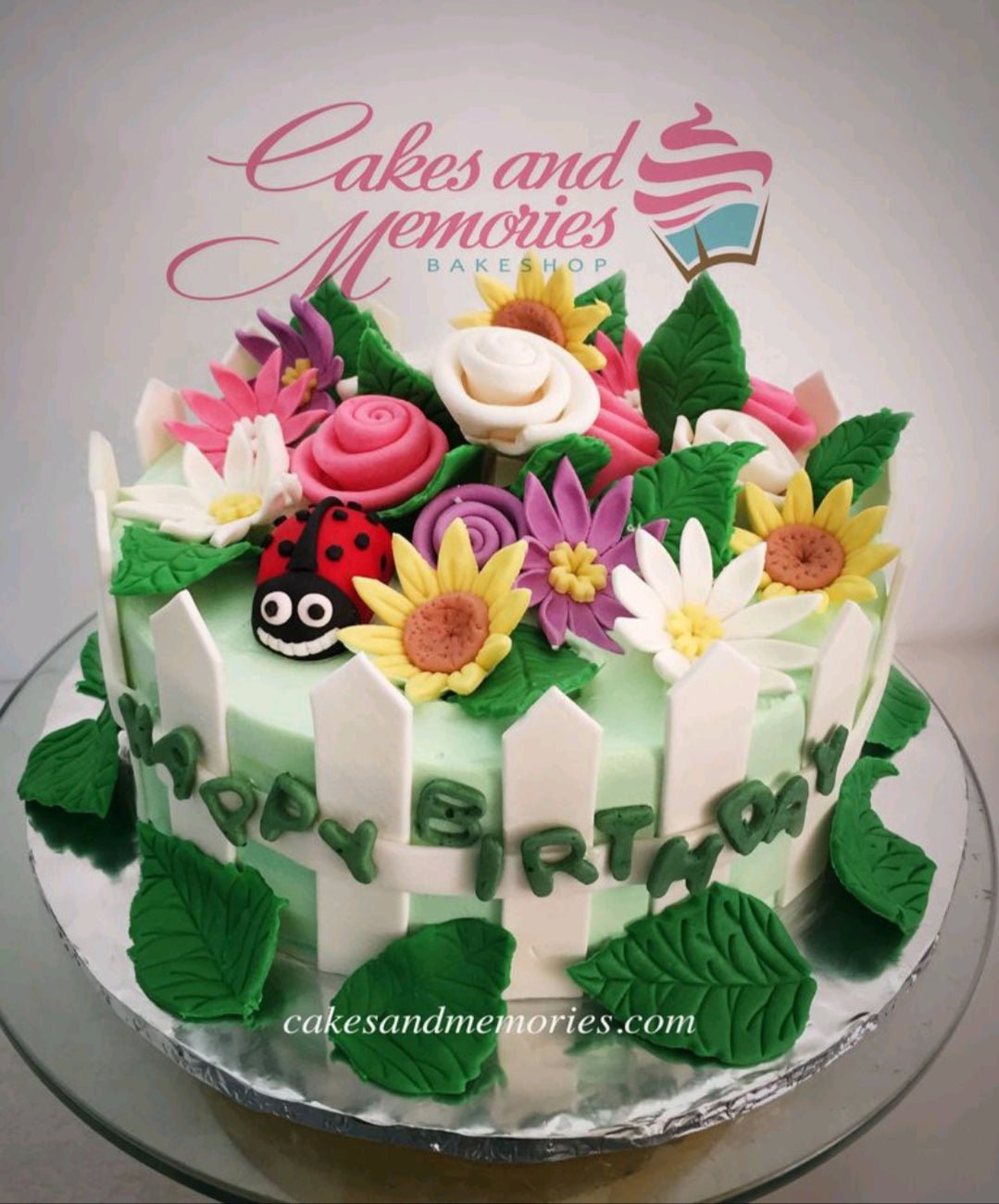 Light green garden themed birthday cake with colorful fondant flowers, a ladybug, and a white picket fence border. Light green garden themed birthday cake with colorful fondant flowers, a ladybug, and a white picket fence border.